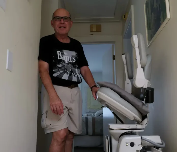 Howard Weinberg, a retired physician living with Parkinson’s disease, stands beside a stair lift at his Virginia Beach home. (Photo by Yiqing Wang/WHRO)